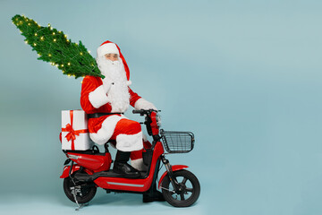 Santa claus on a red electric moped with a gift in the trunk and a Christmas tree on his shoulder...