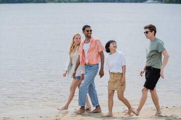 smiling multicultural friends looking at young man while walking along river.