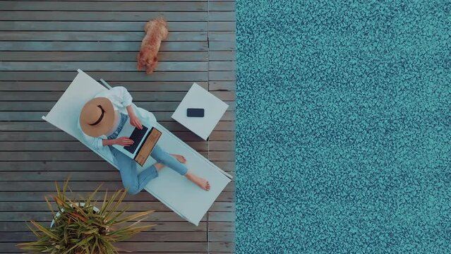 Top down view of a young entrepreneur girl with her brown dog working on her computer in by the resort pool in summer time on a sunbed at sunset. Remote working anywhere