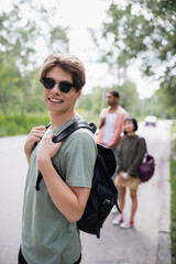 Fototapeta premium smiling traveler in sunglasses looking at camera near blurred interracial friends.