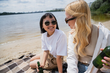 smiling asian woman looking at blonde friend during picnic on beach.