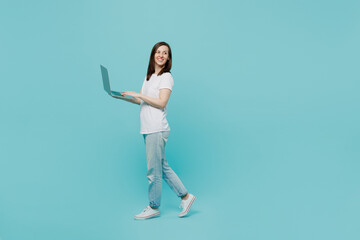 Fototapeta na wymiar Full body side view young smiling happy fun caucasian woman 20s she wear white t-shirt walking going hold use work on laptop pc computer look aside isolated on plain pastel light blue cyan background.