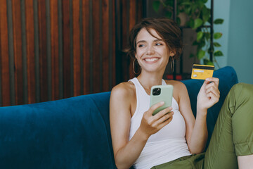 Young happy woman wear white tank shirt using mobile cell phone credit bank card shopping online booking tour sit on blue sofa stay home flat rest relax spend free spare time in living room indoors.