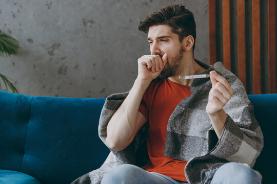 Young sick unhappy ill sad man 20s wears red t-shirt wrapped in plaid hold thermometer caugh covering mouth sit on blue sofa couch stay at home hotel flat spend time in living room indoor grey wall.
