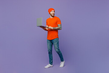 Full body young IT man 20s he wear red hat t-shirt hold use work on laptop pc computer look aside on workspace area isolated on plain pastel light purple background studio. People lifestyle concept.