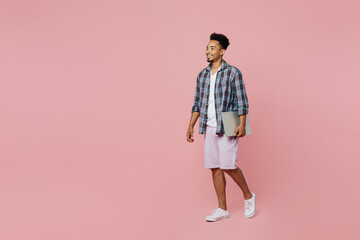 Fototapeta premium Full body side view young smiling student cheerful fun man of African American ethnicity 20s he wear blue shirt hold closed laptop pc computer walk go isolated on plain pastel light pink background.