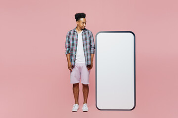 Full body young man of African American ethnicity wear blue shirt stand look at big huge blank screen mobile cell phone copy space mockup area workspace isolated on plain pastel light pink background.
