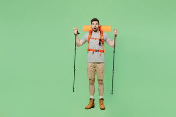 Full body shocked young traveler white man carry backpack stuff mat walk with trakking poles isolated on plain green background. Tourist leads active lifestyle. Hiking trek rest travel trip concept.