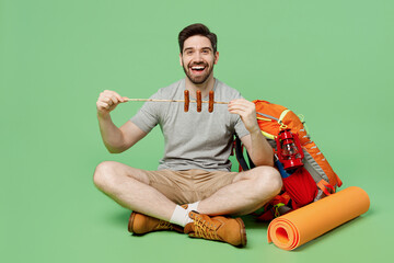 Full body smiling young traveler white man near backpack stuff mat sit roasted sausages on stick...
