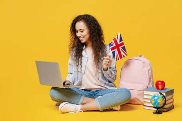 Full body young black teen girl student wear casual clothes backpack bag use work on laptop pc computer hold british flag isolated on plain yellow background. High school university college concept.