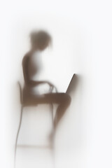 Pretty woman works on laptop computer behind a diffuse white curtain. Body silhouette of a typing...