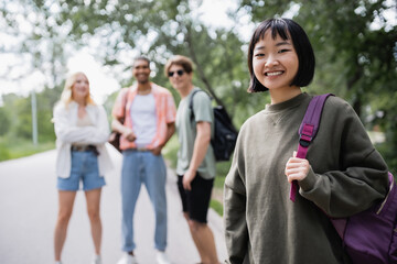 brunette asian woman with backpack looking at camera near blurred multicultural friends.