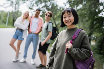 Obraz premium happy asian woman with backpack smiling at camera near blurred interracial friends.