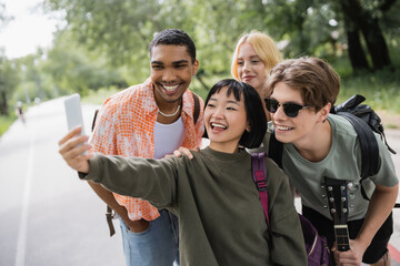 Obraz premium cheerful asian woman taking selfie with multiethnic friends outdoors.