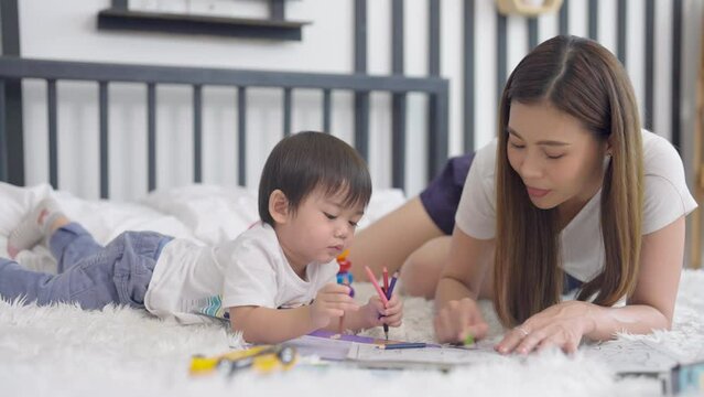 Asian mother and little boy enjoy to paint color with color book on bed in their bedroom together and they look happy fun activities.