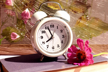  Time to say goodbye to summer: white alarm clock, zinnia flower on a book on a floral background, close-up