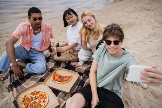 man in sunglasses taking selfie with friends during picnic on riverside.