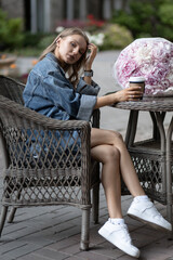 a beautiful caucasian girl in a casual outfit sits in a street cafe next to beautiful peony flowers