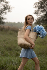 beautiful caucasian girl in a blue dress walks through the field with a basket of flowers