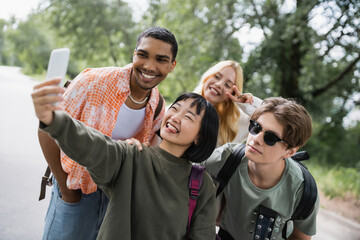 cheerful asian woman sticking out tongue while taking selfie with happy multiethnic friends.