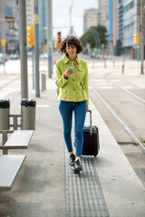 Woman walking with suitcase bag while using smart phone in city