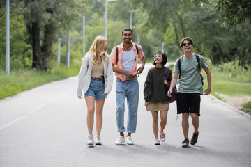 full length of young interracial travelers with backpacks walking along road near forest.