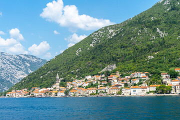 Fototapeta na wymiar Beautiful view of seaside town at foot of mountain on sunny day on coast of sea, houses with terracotta roofs, boats and church in Montenegro, Bay of Kotor, landscape