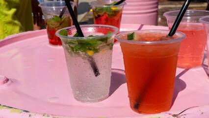 Red and transparent summer cocktails, mojitos and aperol in plastic cups on pink tin barrel on street. Сoncept of picnic, outdoor party