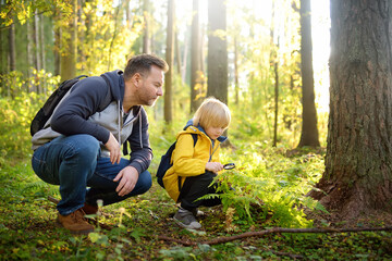 School boy and his father hiking together and exploring nature with magnifying glass. Little boy and dad spend quality family time in sunny forest of second summer. Daddy and little son - obrazy, fototapety, plakaty