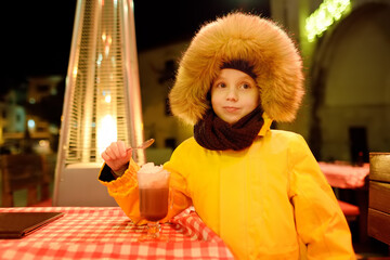 Little boy is drinking hot chocolate with whipped cream in street cafe or restaurant on european...