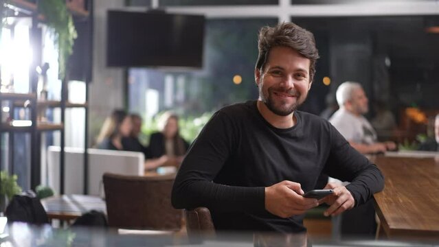 Happy Young Male Customer Sitting At Coffee Shop Holding Phone. Portrait Of A Smiling Man Using Technology Looking At Camera