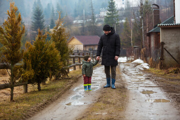 Father and son walking in the fresh air in rubber boots on the puddles and mud after the rain. Little child holding hand of a man