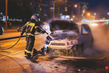 Obraz premium Group of fire men extinguishing and put out burning car crash after road traffic incident, fire fighting operation in the night city, firefighters with fire engine truck vehicle, emergency and rescue