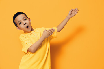 a little boy of preschool age stands on an orange background in a yellow shirt, actively moving his hands pointing to the side, looking at the camera with surprise
