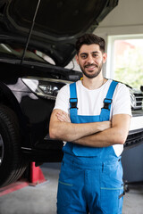 Fototapeta na wymiar Male car mechanic at workplace in spacious repair shop. Portrait of bearded car mechanic crosses hands in a car workshop in blue uniform with equipment looking into camera