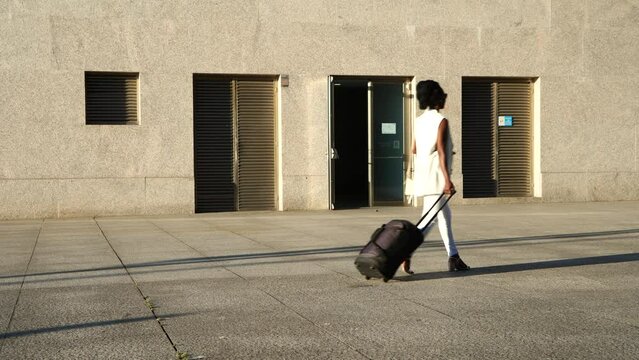 A young businesswoman walking with luggage while holding her phone from left to right