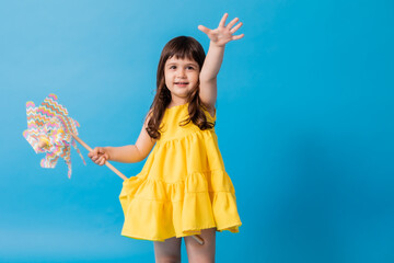 little girl in a yellow dress on a blue background holds a children's toy windmill in her hands