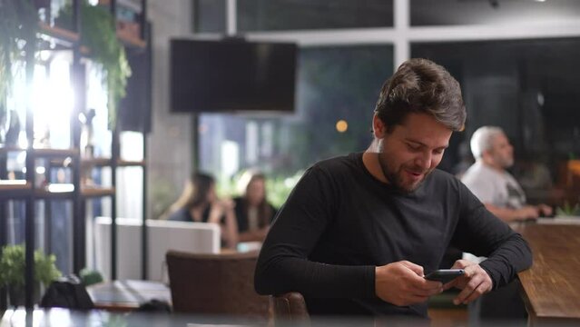 Young man seated inside coffee shop staring at phone screen. Person sitting at cafe store restaurant holding smartphone device