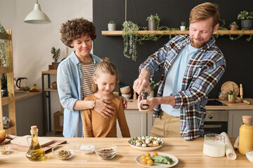 Young man adding pepper in vegetable salad while preparing dinner for his family in kitchen