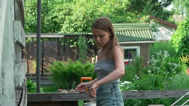 A beautiful caucasian girl in a gray tank top and short denim shorts paints the house with a brush. A European woman paints an old porch with white paint. painting work in a country house.