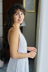 Happy young bride with dark long wavy hair wearing white wedding dress looking at camera while standing in living room at home