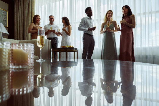 Two Small Groups Of Wedding Guests Or Members Of Bride Family With Champagne Standing In Front Of Table In Living Room At Party