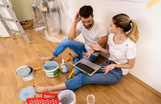 Young couple sitting on the floor choosing color via laptop for painting the wall in their home.