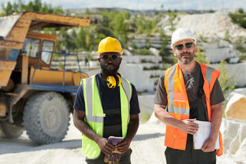 Two multicultural male workers of marble quarry standing in front of camera against dump truck on territory of industrial factory