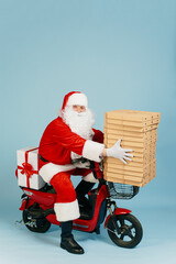 Pizza delivery man in santa claus costume holding a large stack of pizza boxes while sitting behind the wheel of a full length red electric scooter on a blue isolated background with copy space