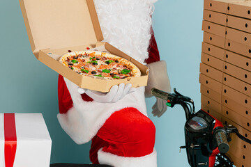 Close-up open box of pizza in the hands of a delivery man in a santa claus costume. He holds in his hands against the background of a red moped with a stack of pizza boxes on a blue studio background