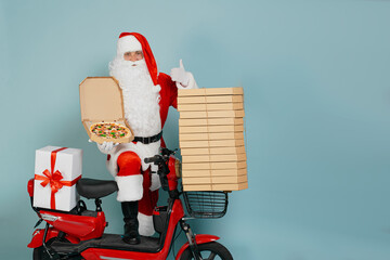 Santa claus demonstrates pizza in an open box close-up on the background of a red moped with a stack of boxes on a blue isolated background with copy space and makes a thumbs up gesture