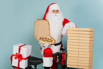 Pizza delivery man dressed as santa claus with an open pizza box in his hand cloaks himself on a stack of boxes on the background of a red scooter and looks at the camera on a blue isolated background