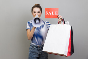 Portrait of woman wearing striped T-shirt announcing shopping discounts and sale, holding shopping...