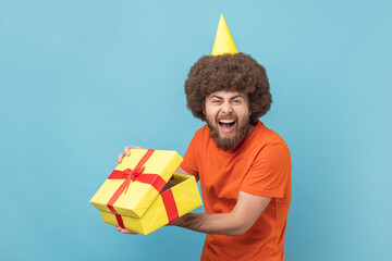 Portrait of happy man with Afro hairstyle wearing orange T-shirt and party cone unpacked present box, looking at camera and laughing. Indoor studio shot isolated on blue background.
